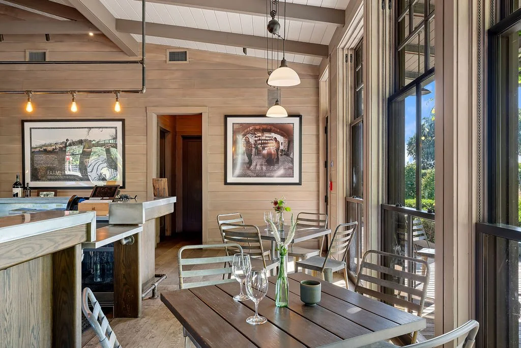 A cozy restaurant or cafe with natural wood decor, large windows, and modern lighting, featuring a small table set with wine glasses and a single flower in a vase.