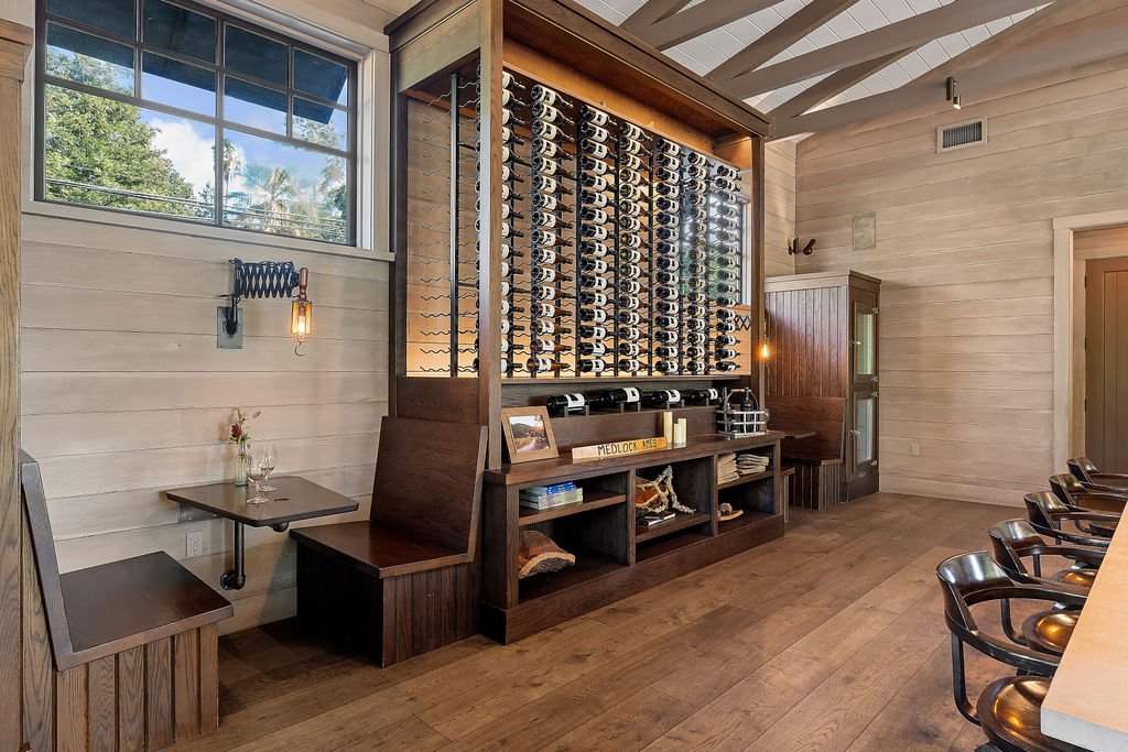 A wine storage rack with bottles on display in a wooden cabinet, situated inside a cozy room with wooden walls and ceiling. There are bar stools along a counter on the right side, and a small table with two chairs and a window on the left side.