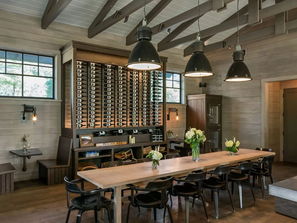 A modern dining room with a long wooden table, black chairs, and three large black pendant lights hanging above. The room features light wood paneling, large windows with natural light, and a wine rack against the wall with neatly arranged bottles an