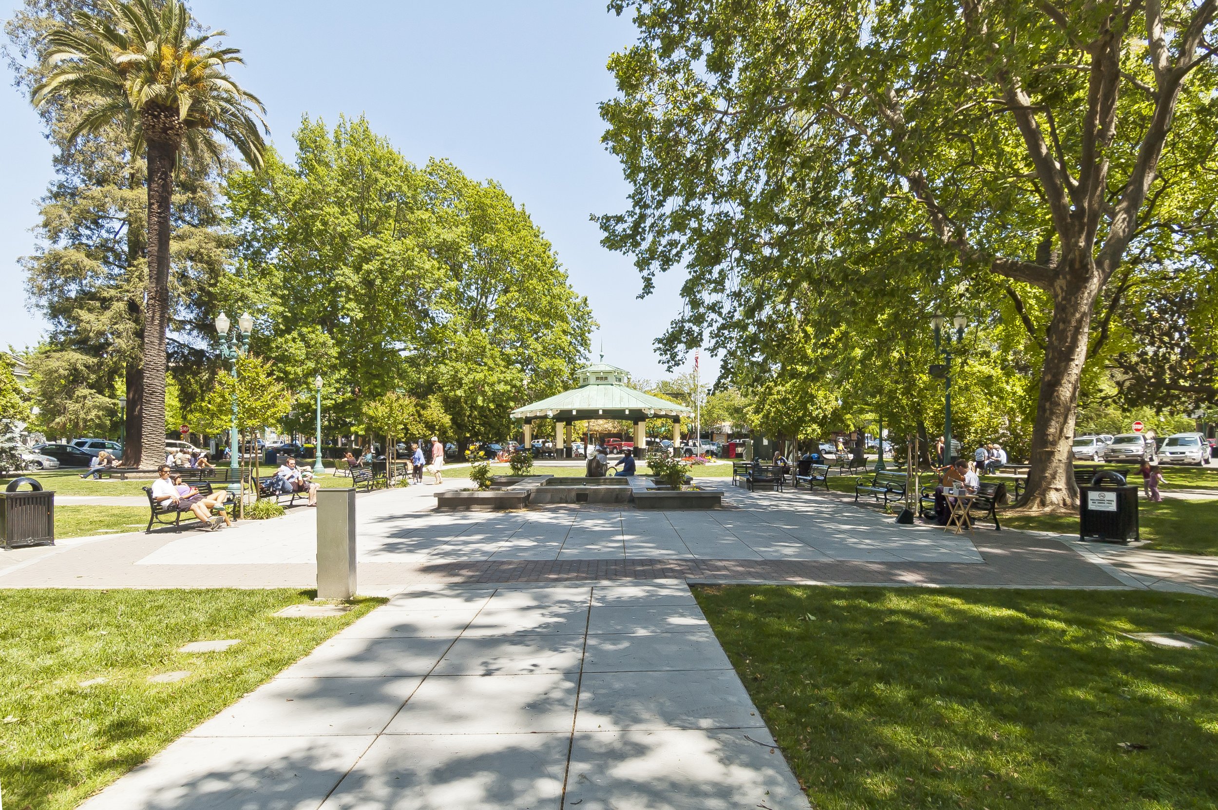 A sunny park with tall trees providing shade, people sitting on benches, and others walking or relaxing. In the background, there is a gazebo with a green roof, surrounded by parked cars and vintage-style lampposts.