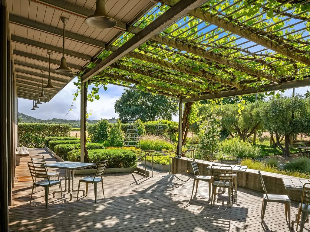 Outdoor patio with tables and chairs, surrounded by lush greenery, trees, and a garden under a partly cloudy sky.