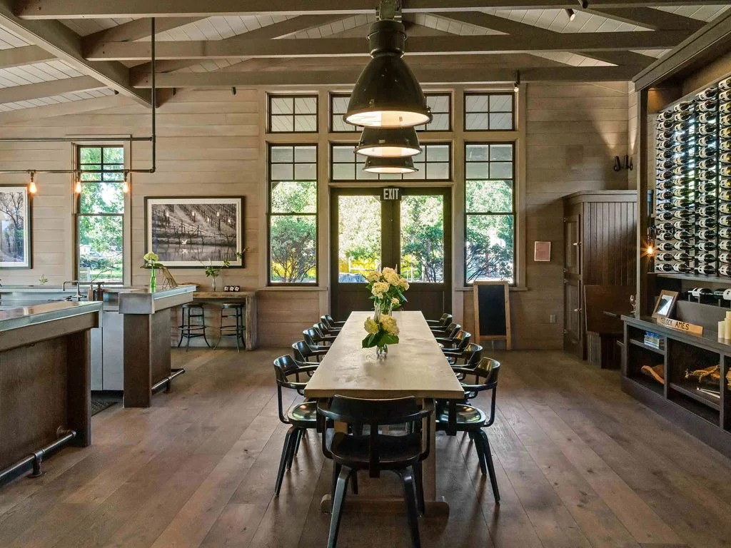 Interior of a cozy dining area with a long wooden table set with flowers, surrounded by black chairs, large windows, a wine rack to the right, and pendant lighting.
