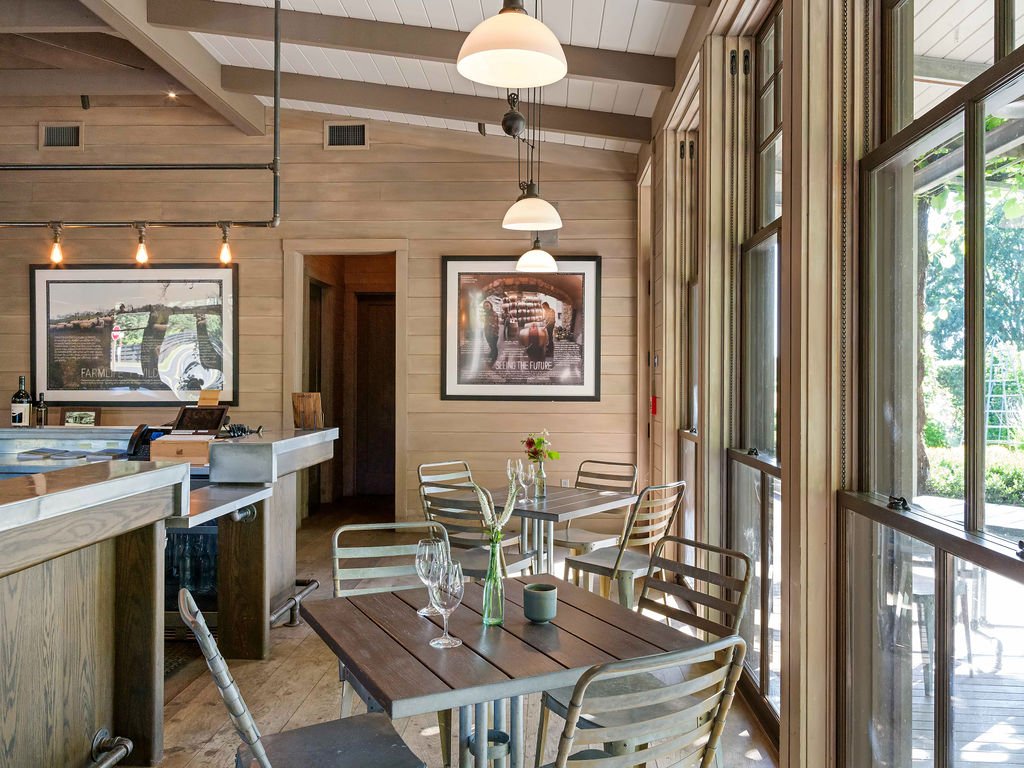 Cozy restaurant interior with wooden walls, large windows, and metal chairs with striped cushions, small tables set with glassware and vases, and framed pictures on the wall.