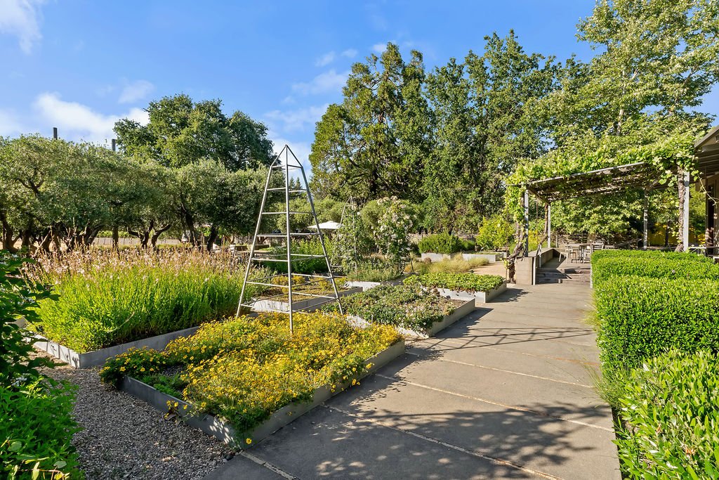 A well-maintained garden with raised flower beds, a metal ladder, trees, and a paved walkway under a partly cloudy sky.