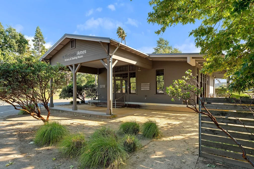 Exterior of a winery tasting room building with trees and bushes in front, clear blue sky, and a sign that reads 'Metropolitan Ames Winery & Tasting Room'.
