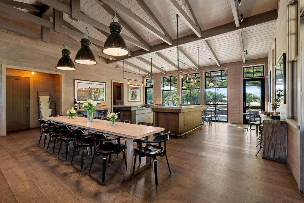 Interior of a modern dining and kitchen area with wooden floors, large windows, and high ceilings. Features a long dining table with chairs, pendant lights, and a kitchen island with seating. Decor includes framed art and potted plants.