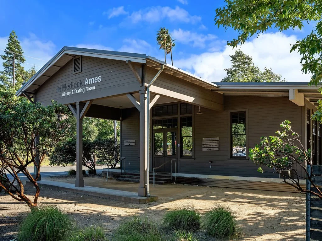 Exterior of a gray building labeled 'Medlock Ames Winery & Tasting Room' with trees and bushes around, under a blue sky with some clouds.