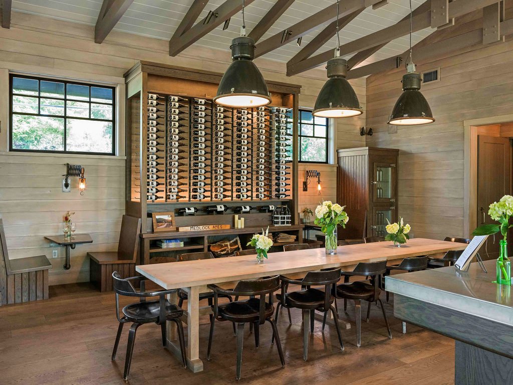 A cozy taproom with a long wooden table decorated with vases of white flowers, surrounded by black chairs. There are three large black pendant lights hanging above the table. A wall of wine bottles is behind the table. The room has wooden walls, large windows, and a small seating nook with a table to the left.