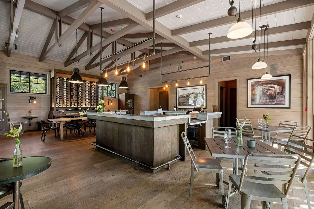 Empty restaurant interior with wooden walls, tables, and chairs, a bar with pendant lights, and artwork on the walls.