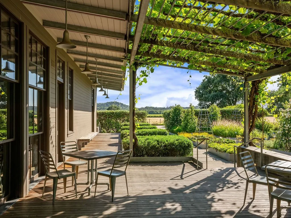 Outdoor patio with wooden flooring, metal chairs, and tables under a pergola with leafy vines. Garden and open fields in the background.