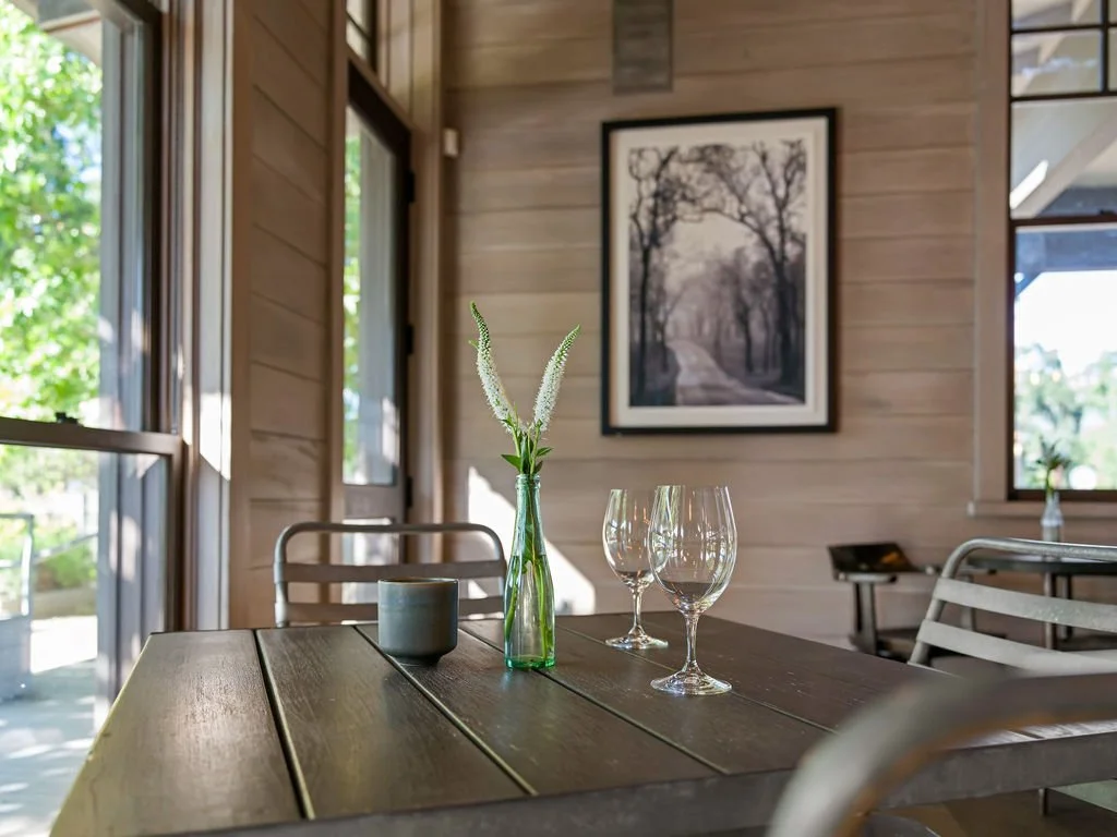 A wooden dining table with a small green vase containing white flowers, two empty wine glasses, and a gray cup, inside a room with wood-paneled walls, large windows, and a framed black-and-white photograph of a tree-lined path.