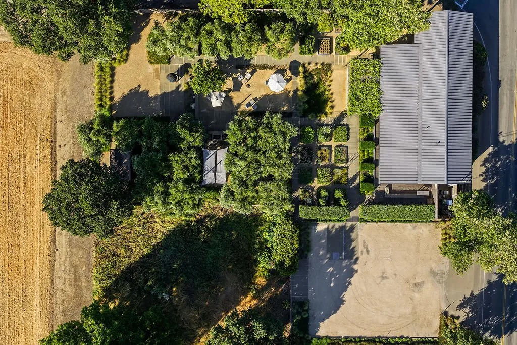Aerial view of a house with a metal roof, surrounded by trees, garden beds, and a patio with umbrellas, near a tennis court and a baseball field.