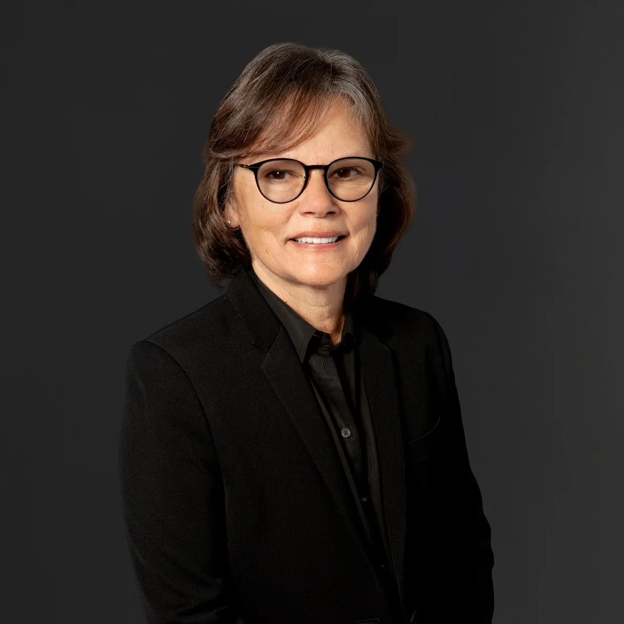 A professional woman with short brown hair and glasses, wearing a black suit and shirt, smiling against a dark gray background.