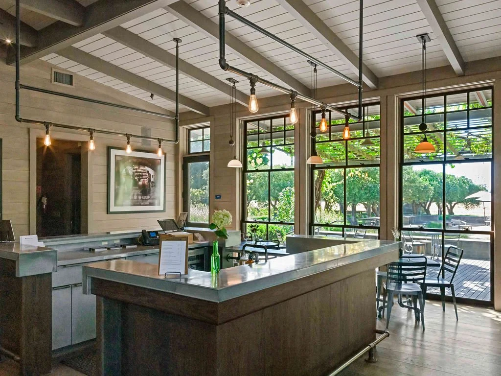 Bright restaurant interior with large windows, wooden ceiling beams, pendant lights, and a marble counter, overlooking outdoor trees and patio.