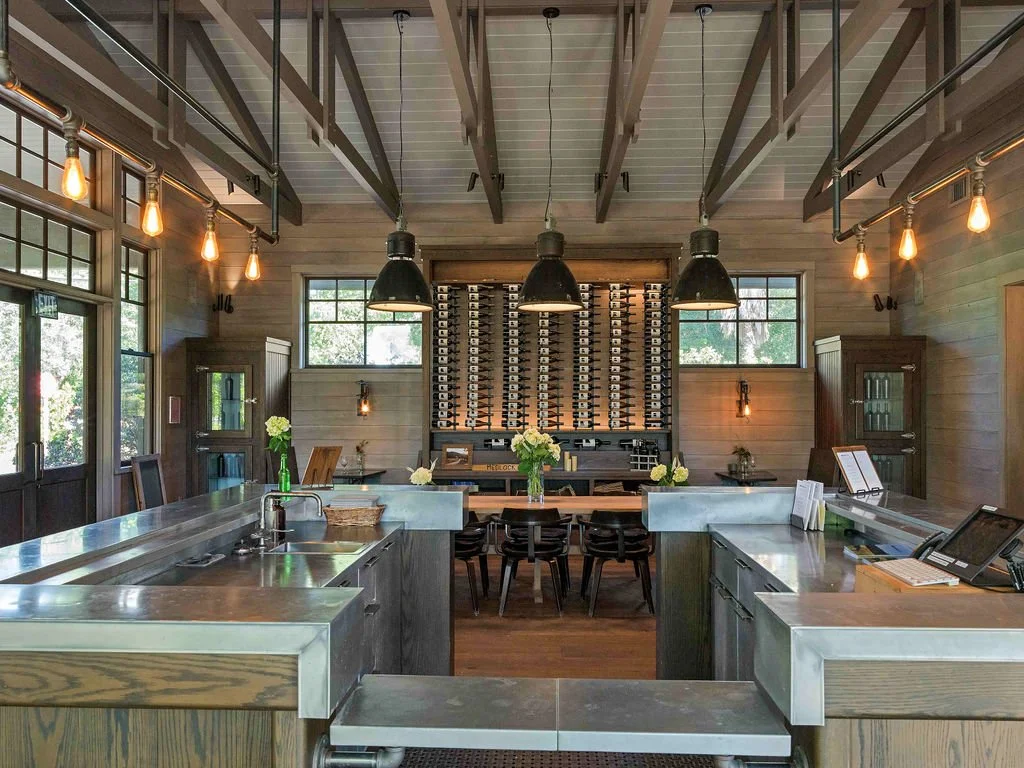 Interior of a modern wine tasting room with a metal bar, black chairs, wine bottle racks, and warm lighting
