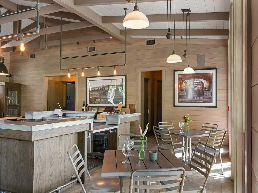 A cozy restaurant or cafe with wooden interior, white hanging pendant lights, and a bar counter. There are small tables with wine glasses, a plant, and framed pictures on the walls. Soft natural light fills the space.