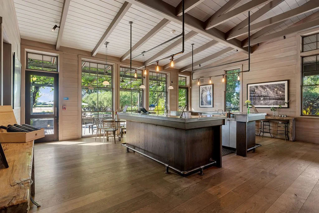 Bright, airy cafe interior with wooden floors, large windows, a bar counter, hanging pendant lights, and outdoor seating visible through the windows.