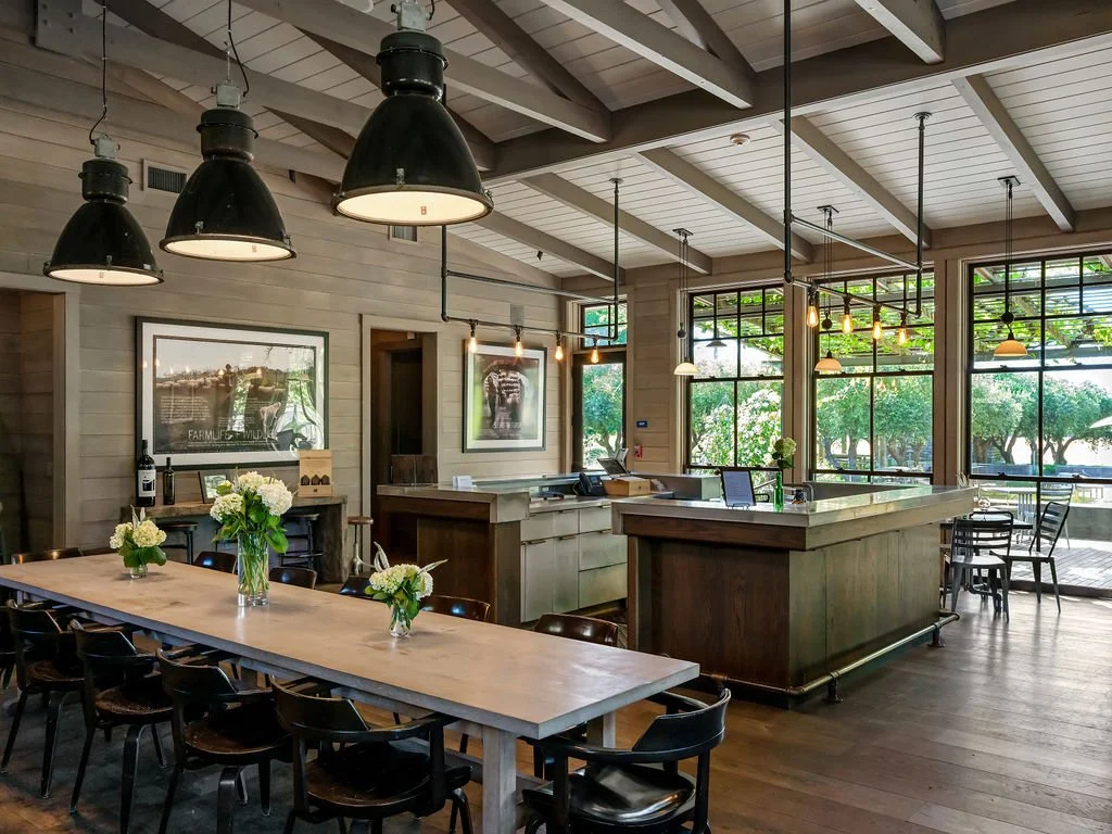 Interior of a modern restaurant with a large wooden dining table, black chairs, and vases of white flowers. There are hanging industrial-style lamps and large windows showing greenery outside.