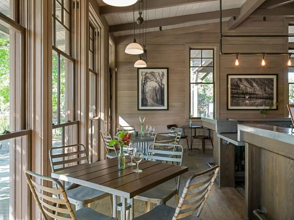 A cozy dining area with wooden walls and large windows, featuring a wooden table with metal chairs, a small vase with flowers, and framed black-and-white landscape photographs on the wall.