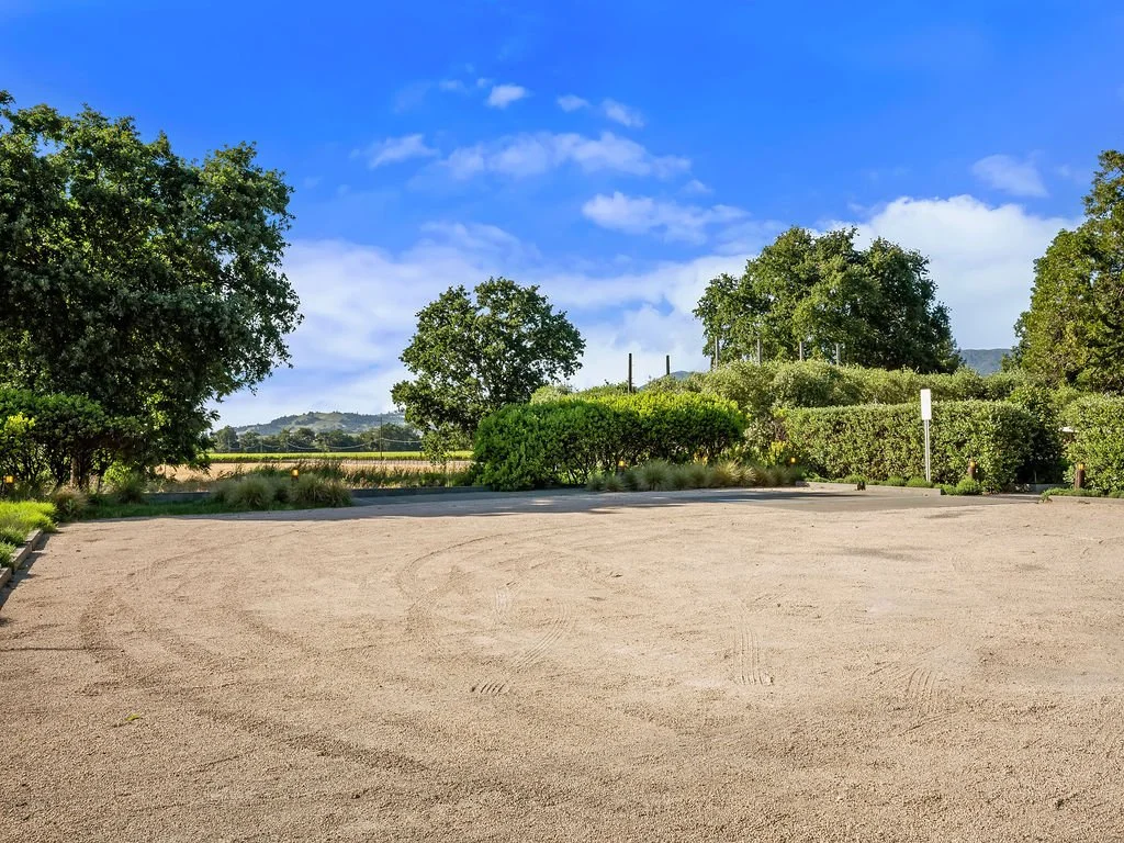 An empty, sandy parking lot surrounded by green bushes and trees against a bright blue sky with some clouds.