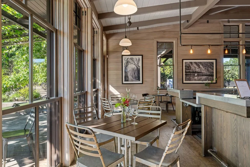 Interior of a cozy restaurant with wooden walls, large windows, hanging pendant lights, and framed black-and-white photography, with tables set for dining.
