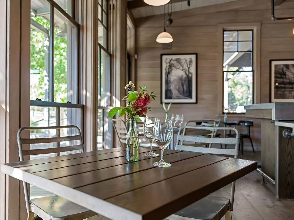 A wooden dining table set with a small flower arrangement and two wine glasses inside a bright, cozy restaurant with wooden walls and framed artwork.