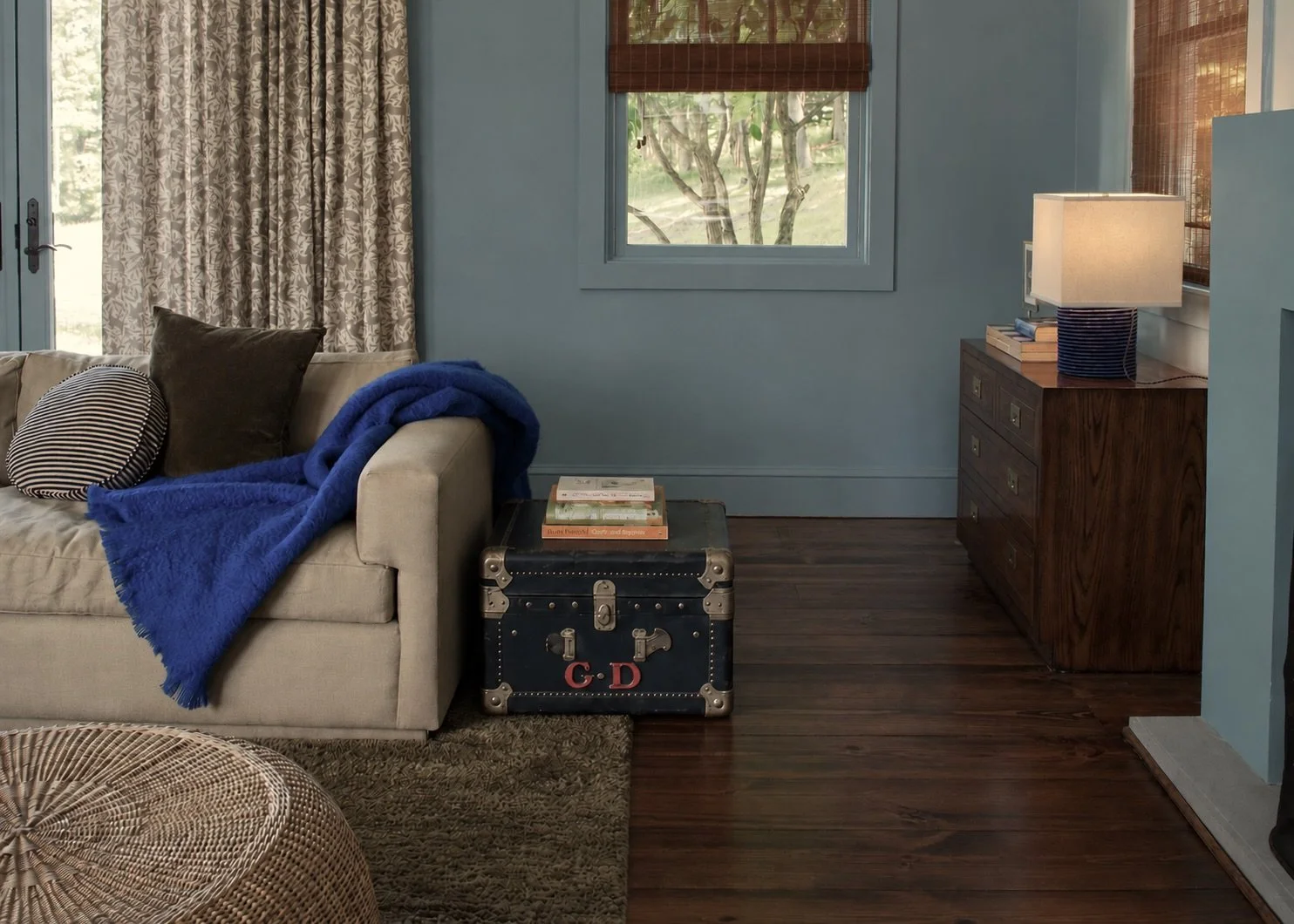 Blue-walled living room corner with a linen sofa, a cobalt wool throw, a studded campaign trunk and a walnut chest beneath a shuttered window
