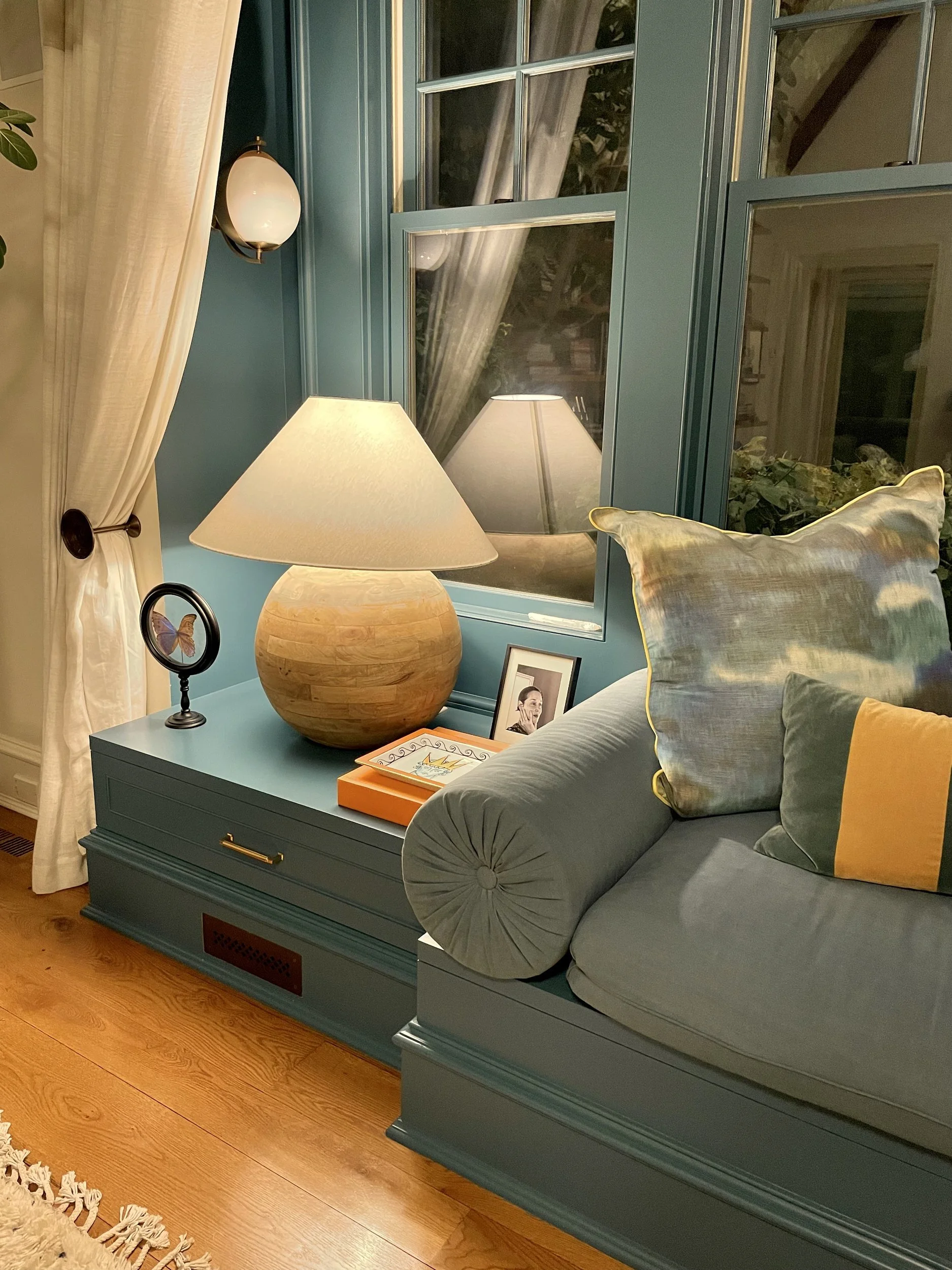 Window seat detail with teal panelling and drawers, a turned wood sphere lamp, blue bolster and cushions, and a brass globe sconce