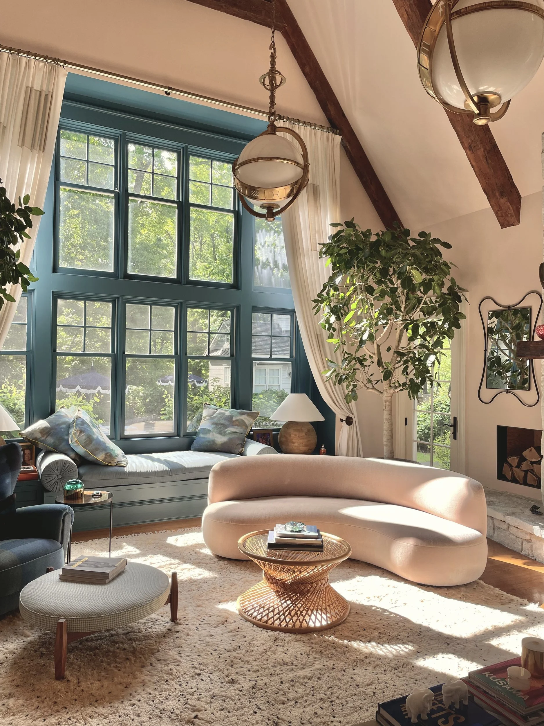 Living room with a vaulted beamed ceiling, a teal-painted window wall, a curved cream sofa, rattan drum table and brass globe pendants