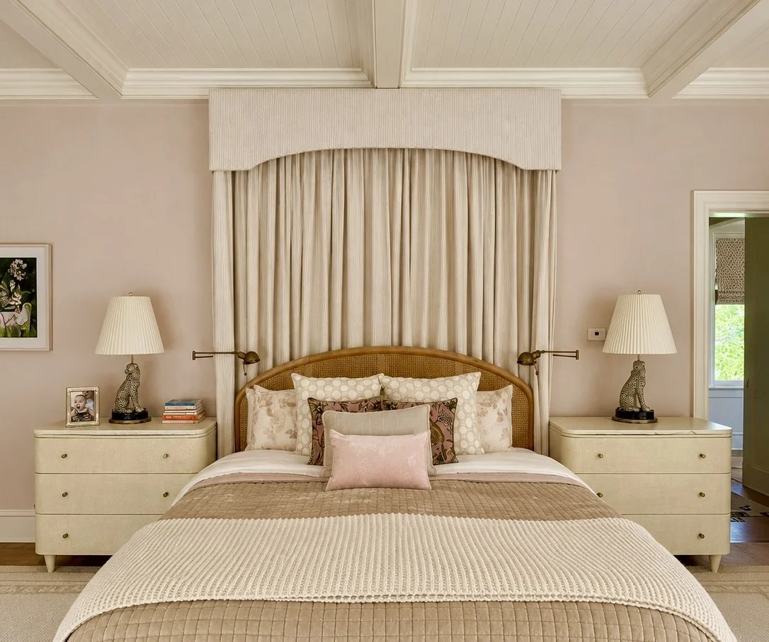 Bedroom with pale pink walls, a pleated canopy over a caned headboard, matching cream chests and layered beige quilted bedding