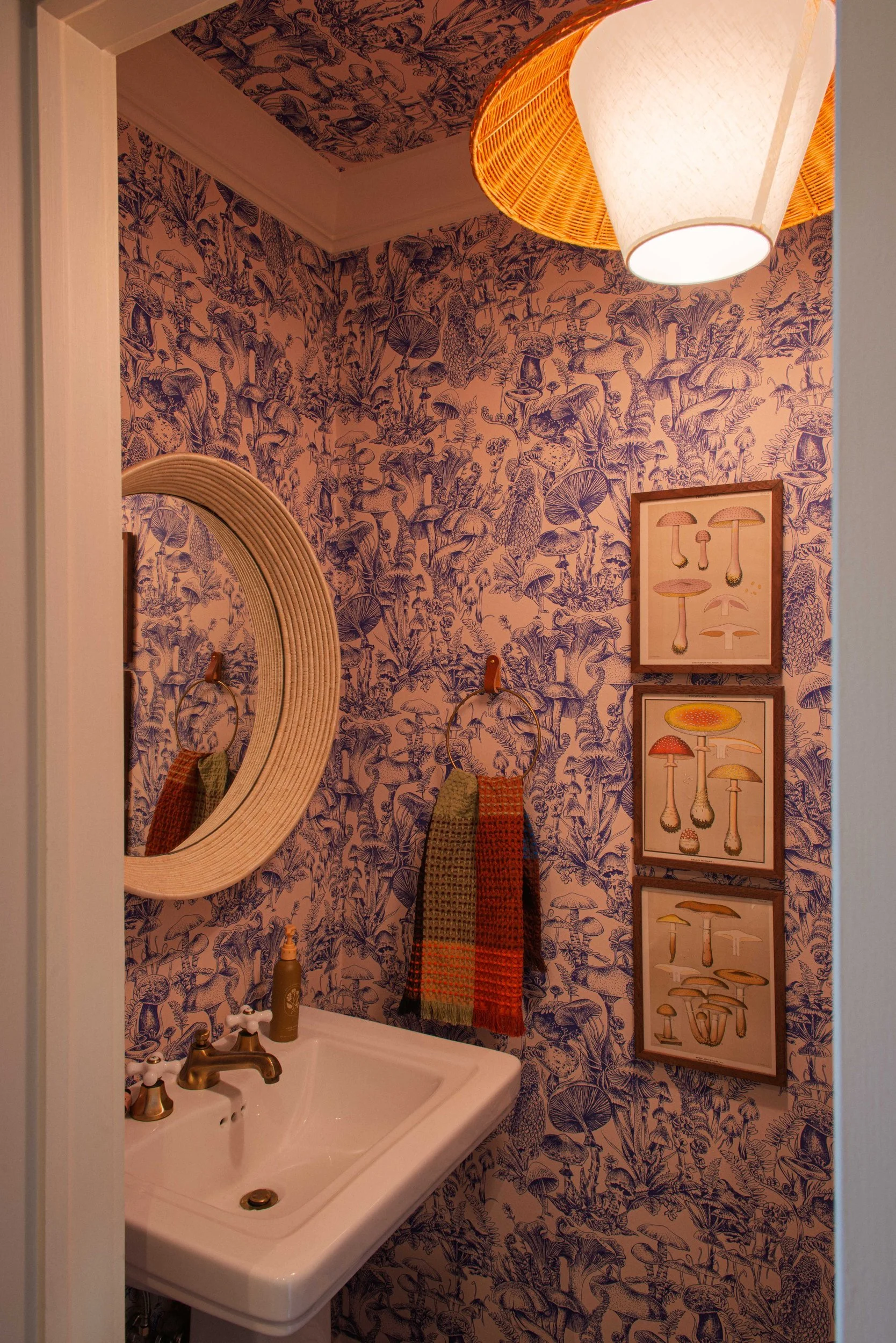 Powder room papered in a blue mushroom print with a pedestal sink, brass taps, a rattan-framed mirror and framed botanical prints