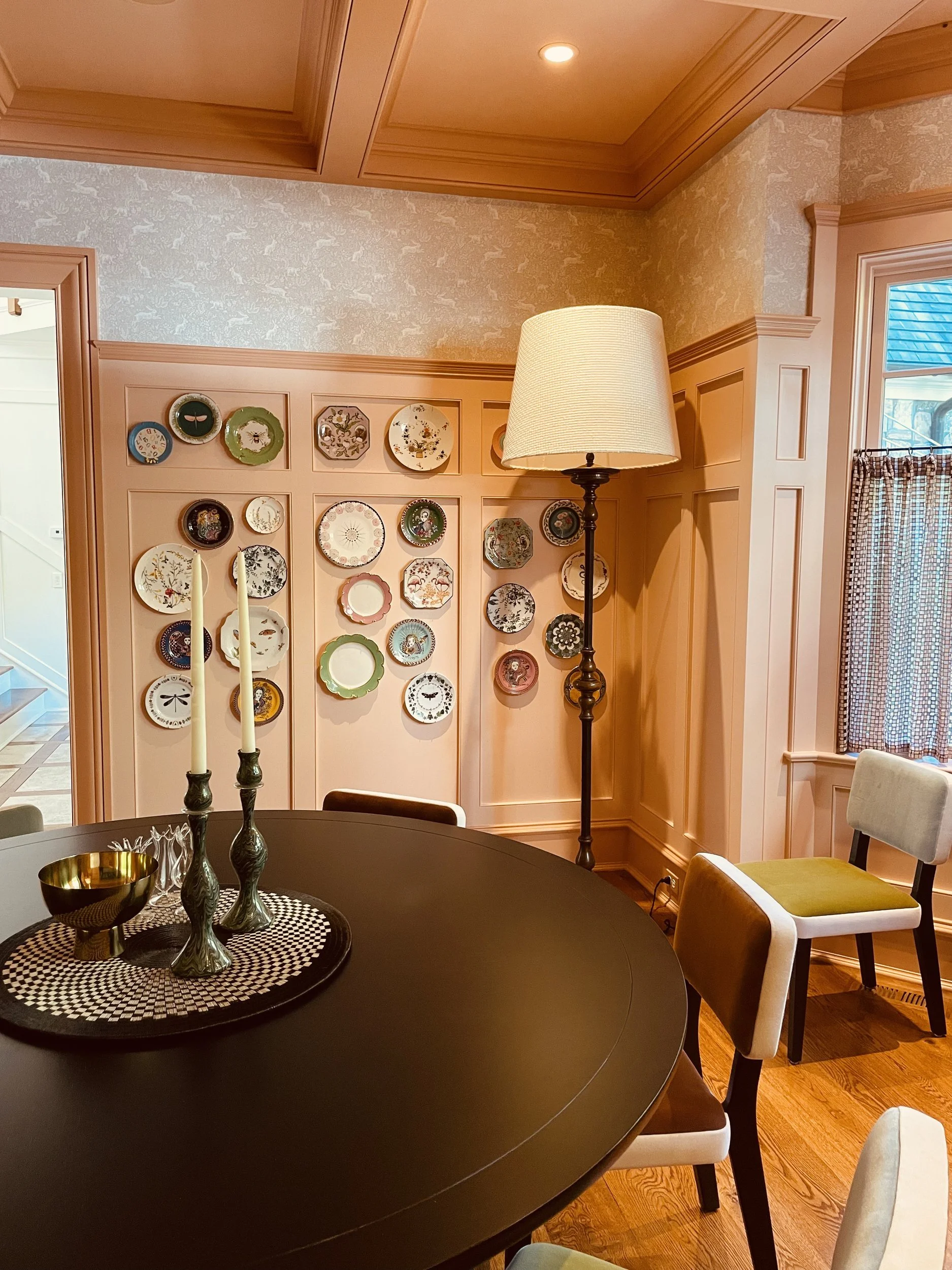 Dining room with peach panelling, a wall of mixed patterned plates, a dark round table and olive upholstered chairs