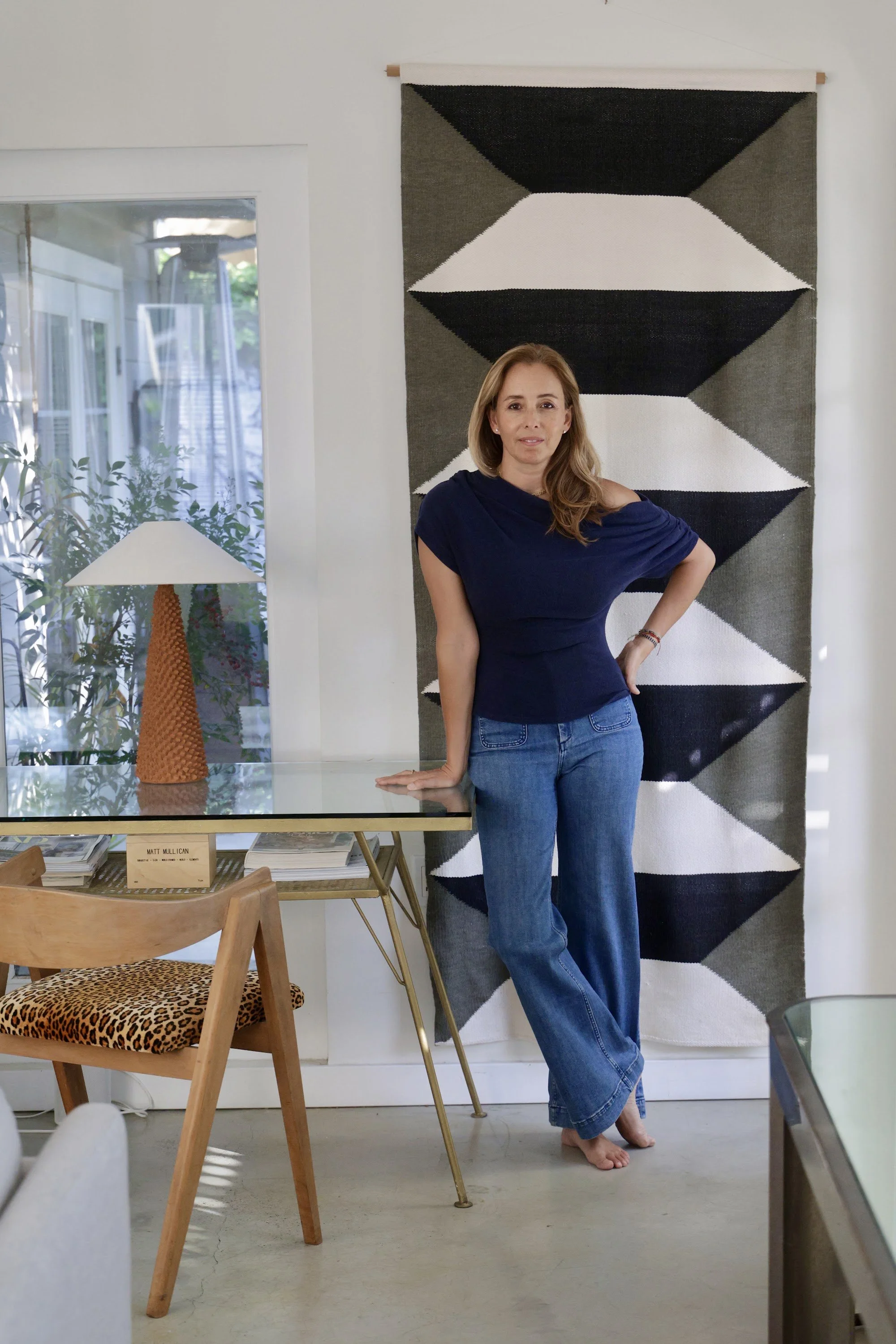 Interior designer Melissa Pinto standing in a room she designed, beside a glass-topped desk and a graphic black and white wall hanging