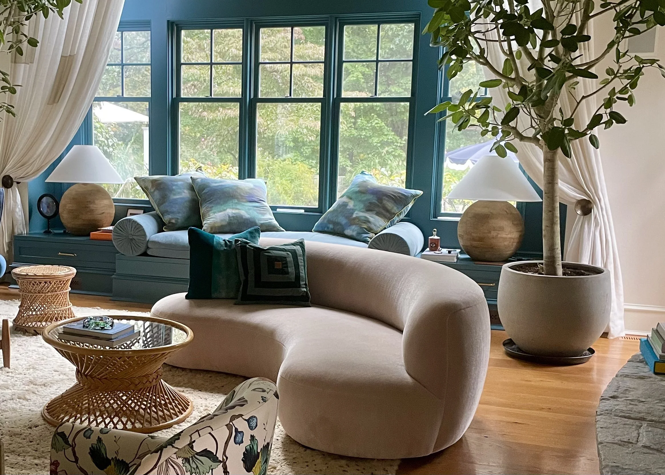 Living room with teal window-seat joinery, a curved cream sofa, woven rattan side tables, a potted tree and a floral armchair