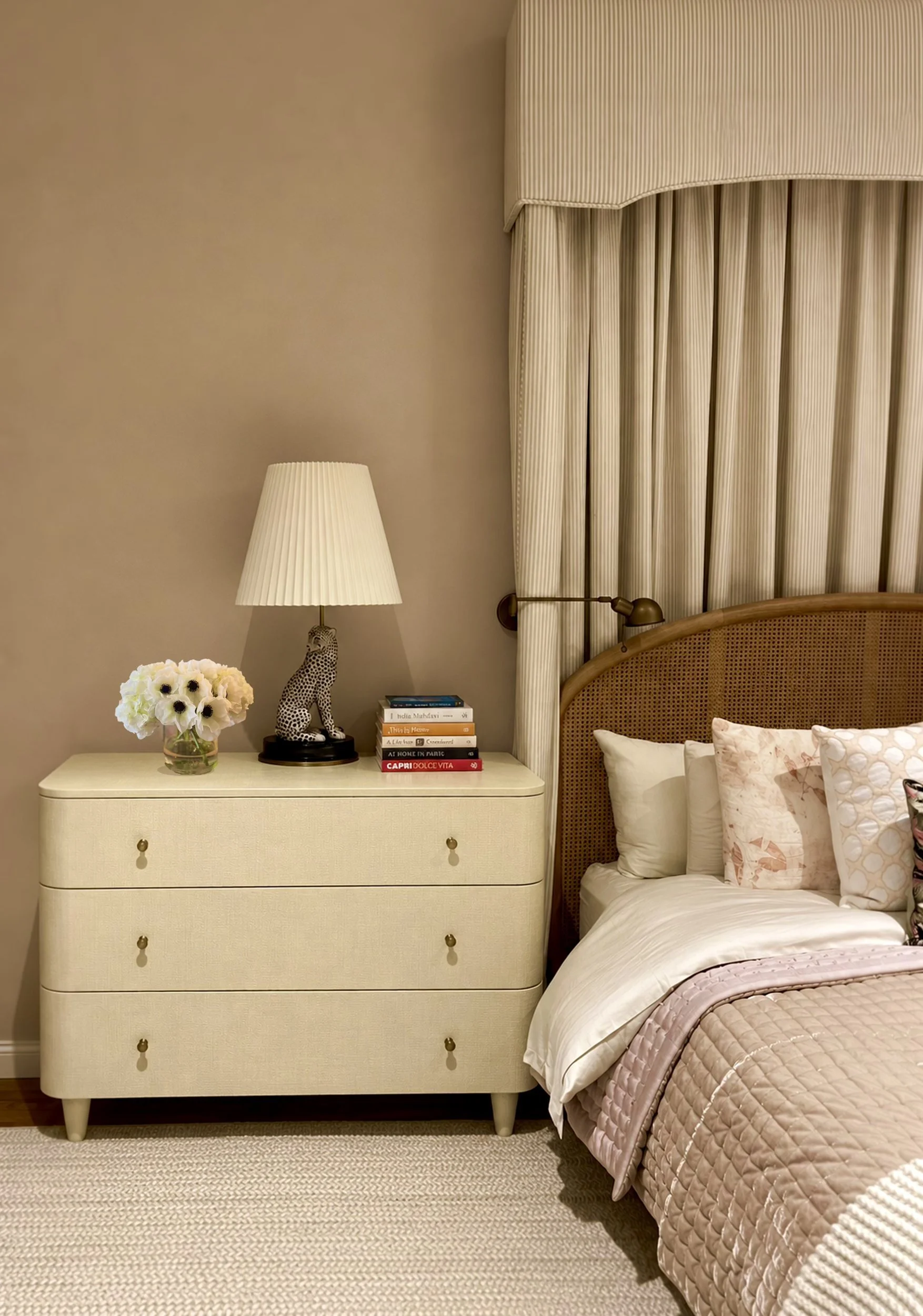 Bedroom detail with a cream linen-wrapped chest, a pleated lamp on a leopard base, a caned headboard and blush quilted bedding