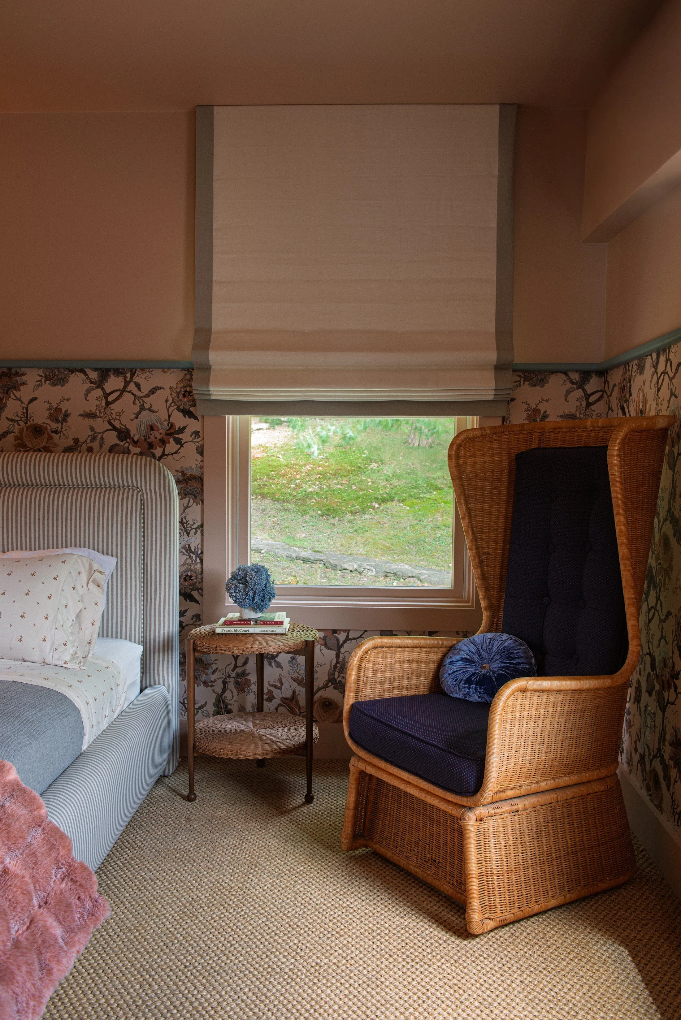 Bedroom with floral wallpaper, a wicker wingback chair and a window overlooking the garden