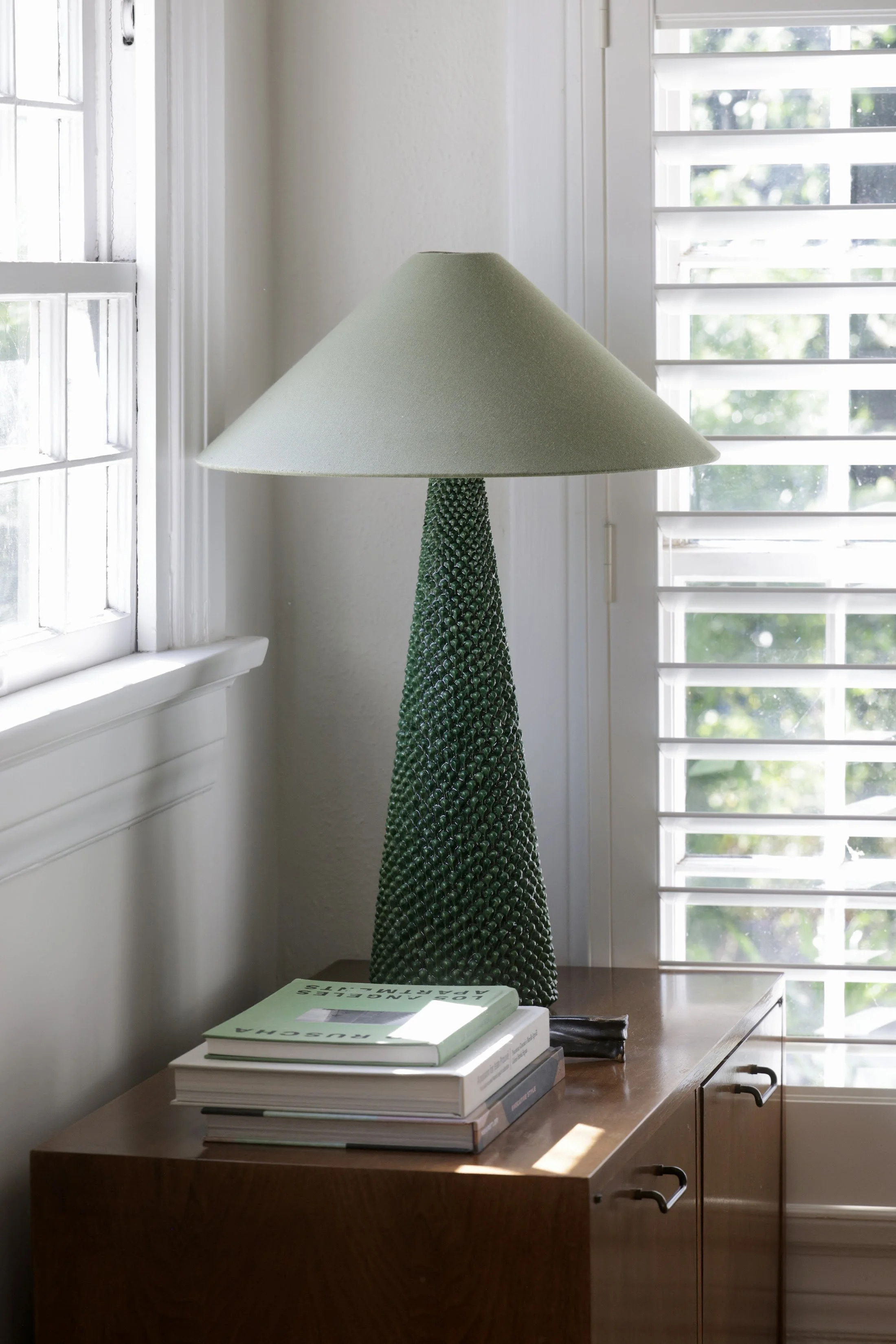Green textured table lamp on a walnut desk beside a shuttered window