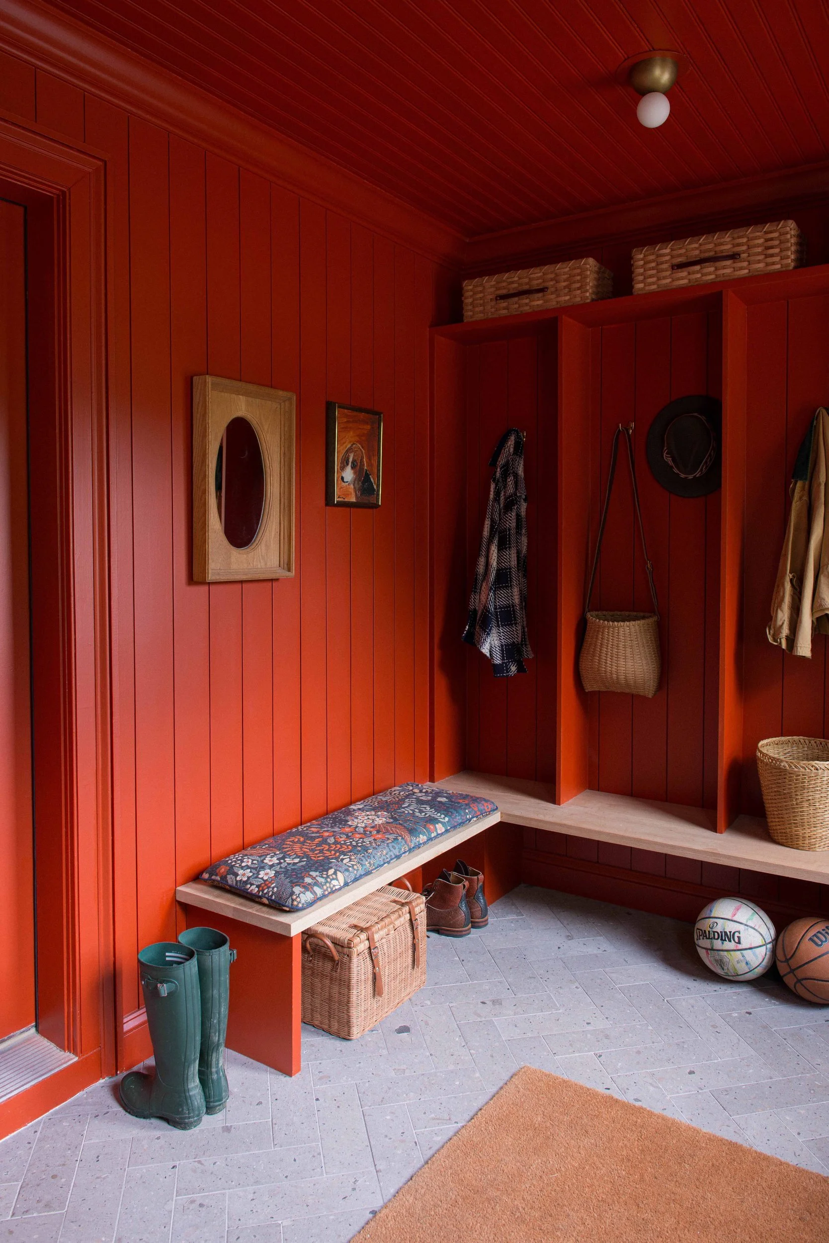 Mudroom panelled in orange-red with open lockers, an oak bench, woven baskets and a herringbone stone floor