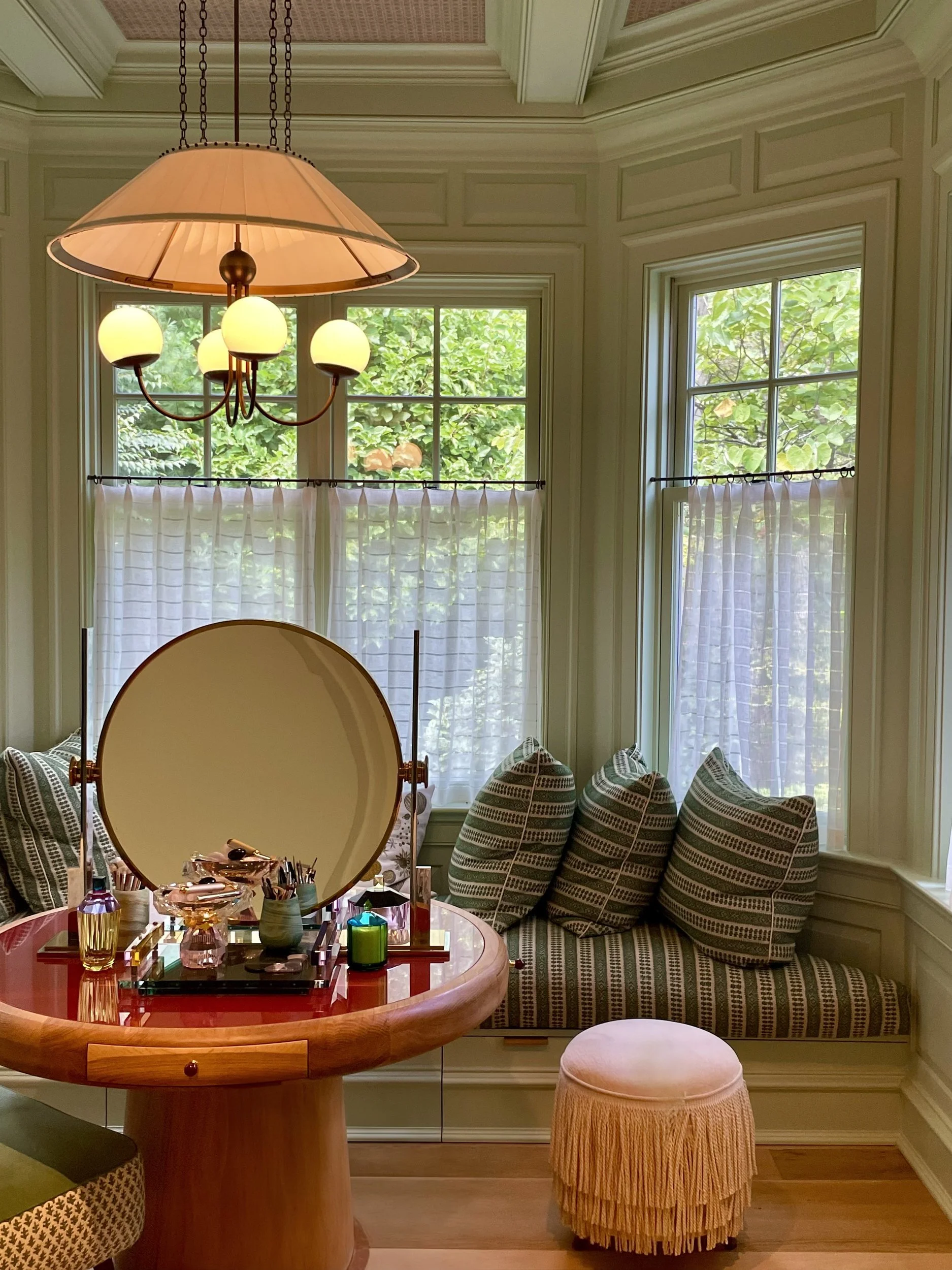 Dressing corner in a bay window with a red lacquer and wood vanity, round mirror, green striped window seat and fringed stool
