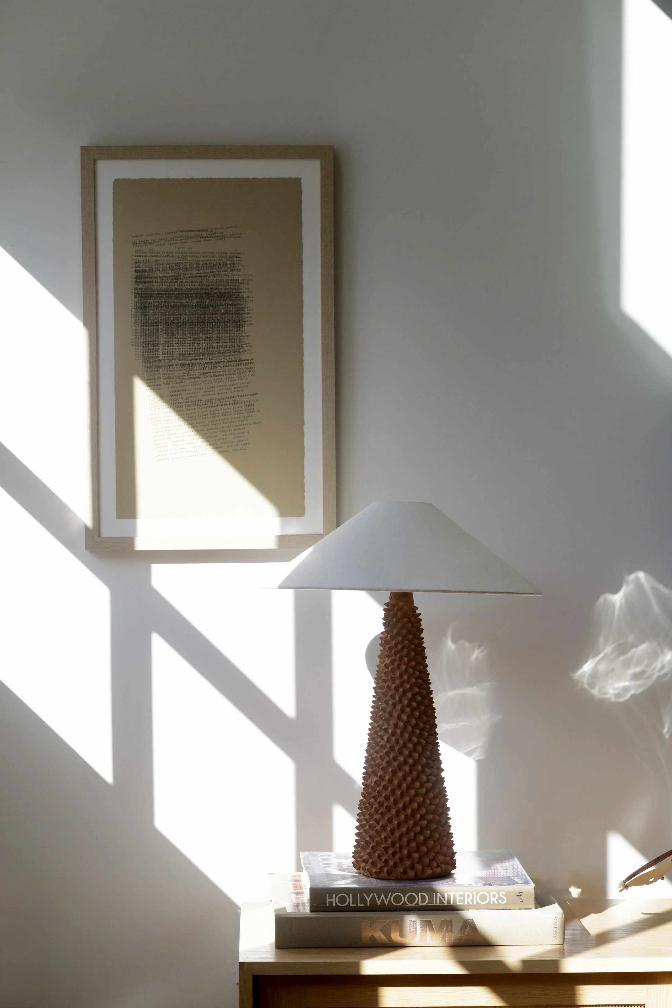 Sunlit corner with a tapered terracotta lamp, stacked books and a framed drawing
