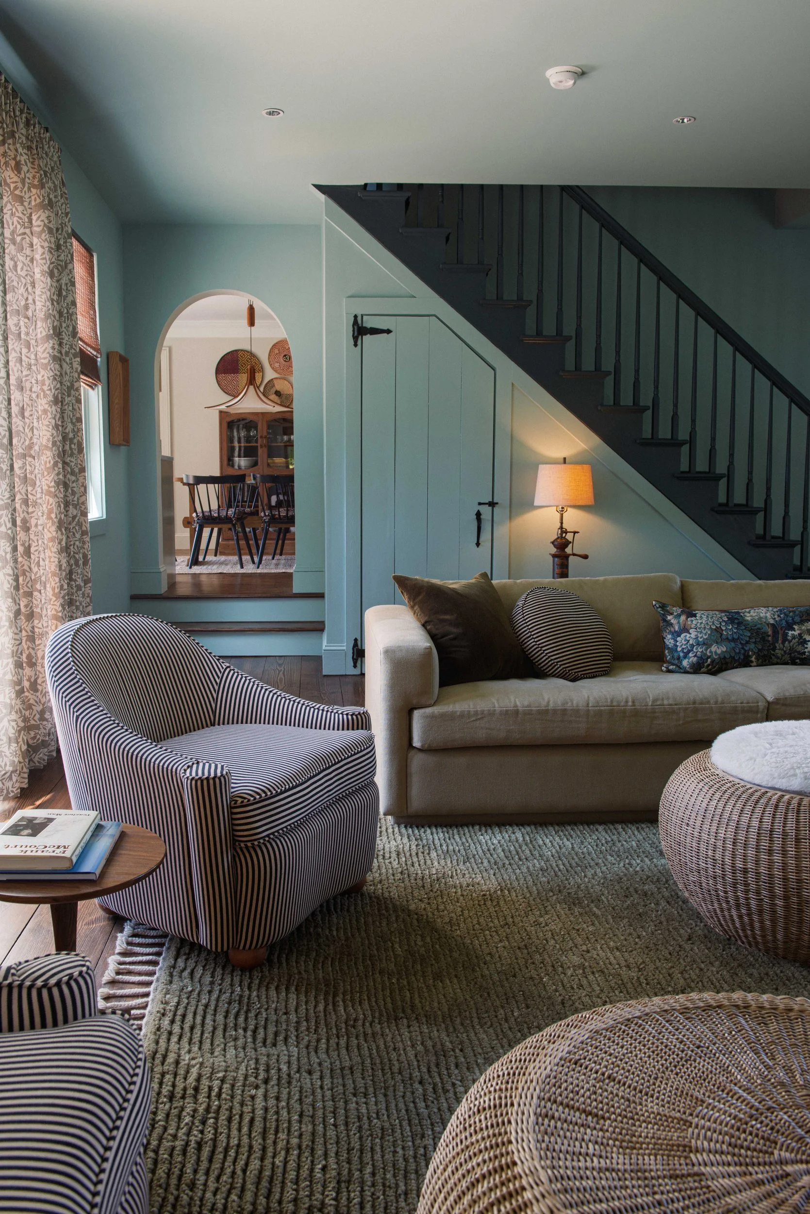 Living room with pale blue-green walls, a navy striped armchair, a linen sofa and a black-painted staircase beside an arched opening to the dining room