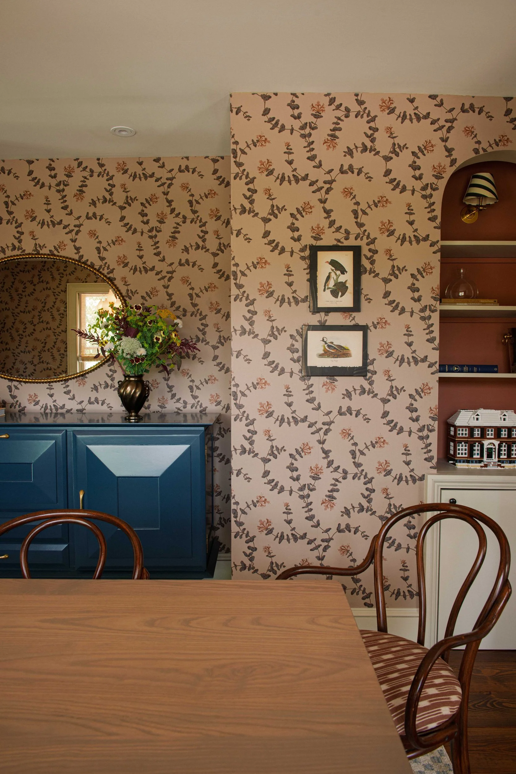 Dining room with honeysuckle wallpaper, a petrol blue sideboard, bentwood chairs and a terracotta-painted built-in bookcase