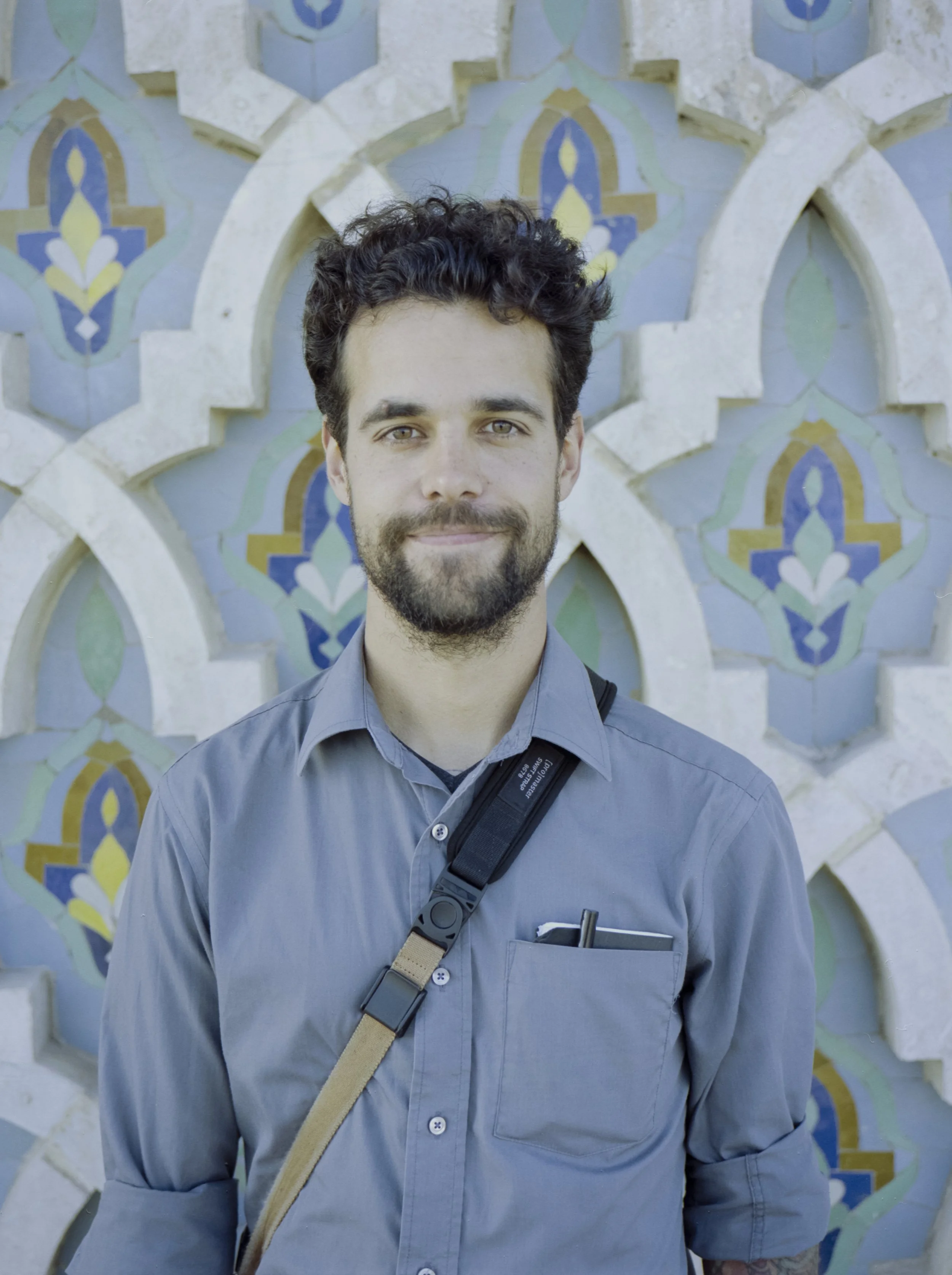 A smiling man with dark, curly hair and a beard stands in front of a decorative wall with colorful mosaic tiles in patterned arches.