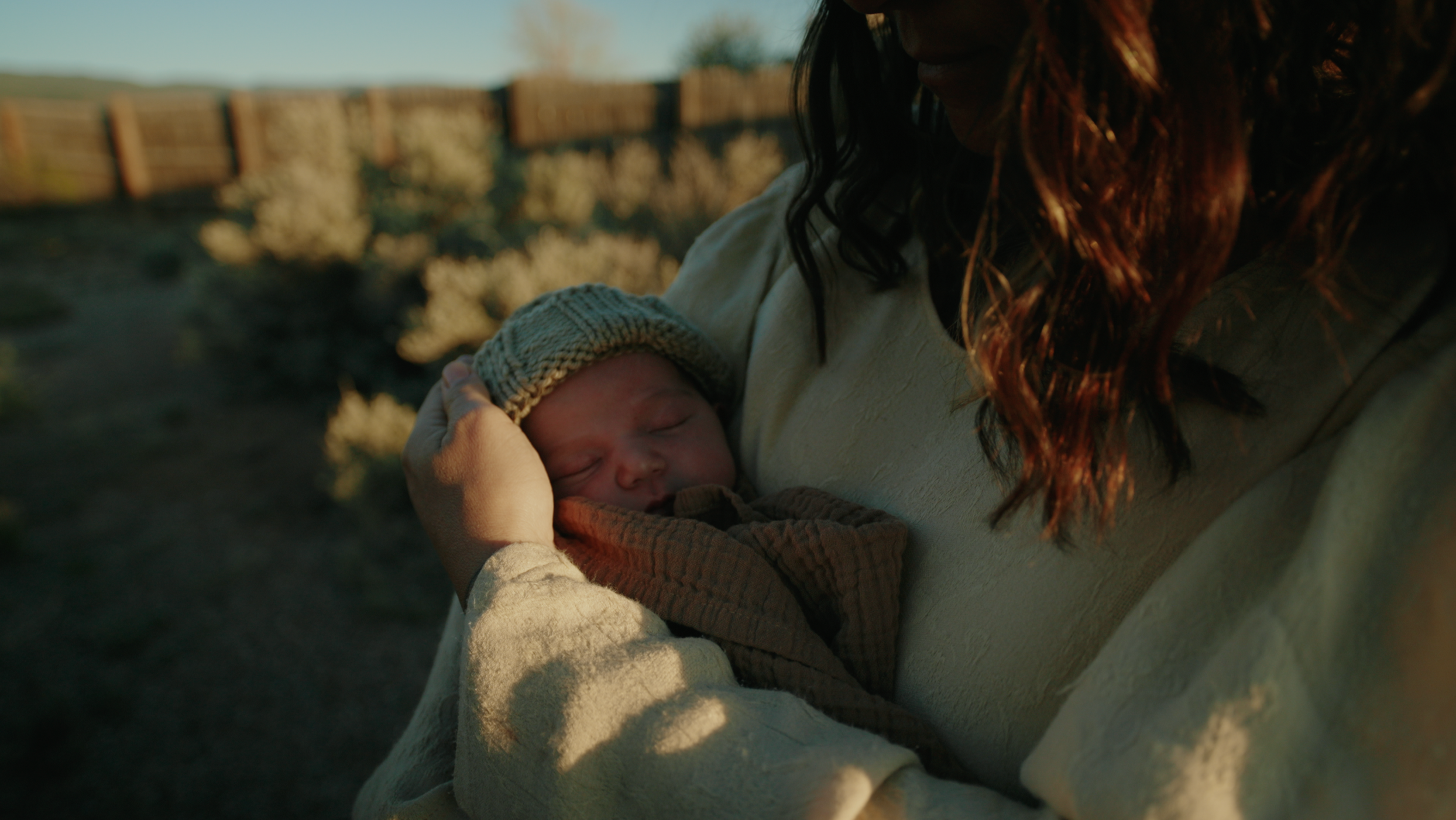 A woman with dark, wavy hair holding a sleeping newborn baby outdoors during sunset.