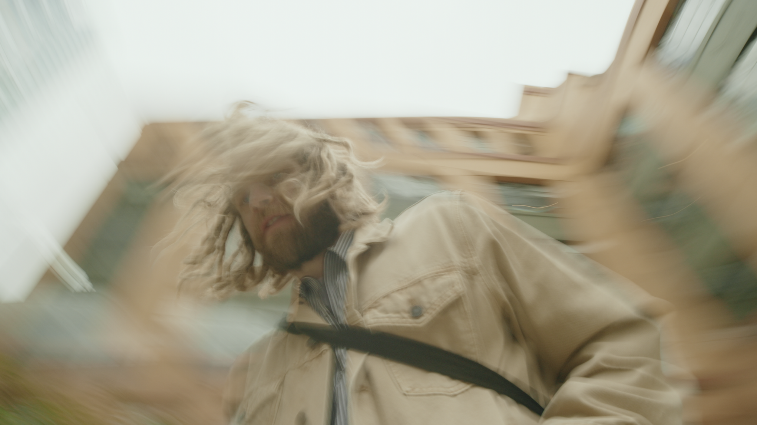 Blurred image of a man with long, curly blonde hair and a beard, wearing a beige jacket and a striped shirt, looking down with a background that appears to be a room with wooden furniture.
