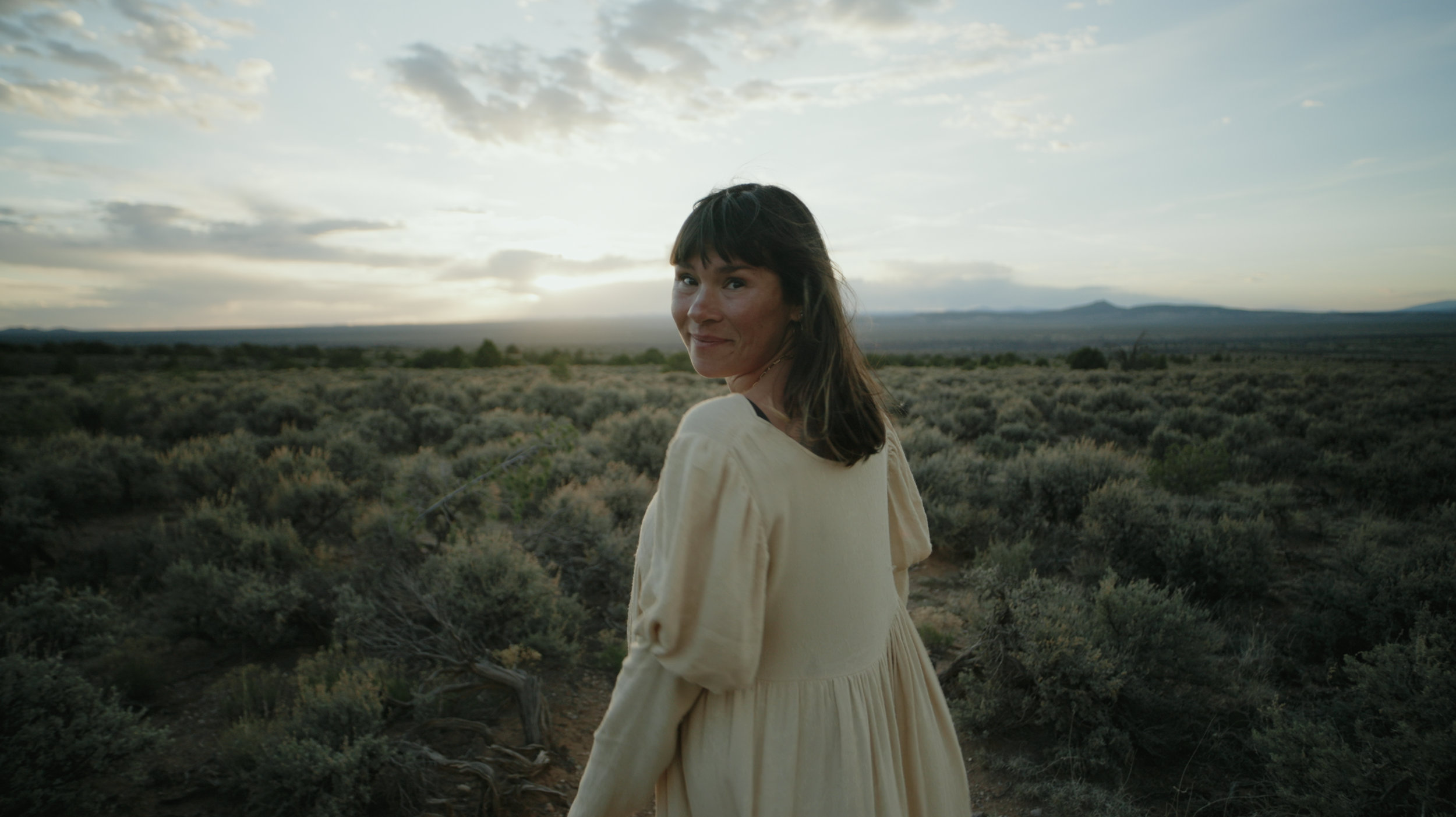 Woman with dark hair wearing a beige dress, standing in a desert landscape at sunset with cloudy sky and mountains in the distance.