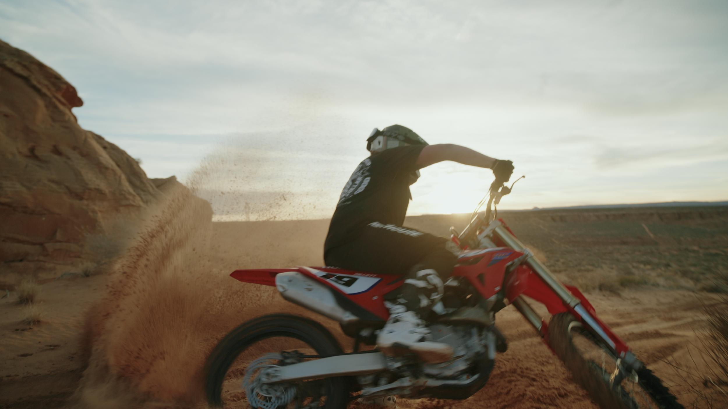 A person riding a red dirt bike on a trail in a desert landscape during sunset, kicking up dust as they speed past a rock formation.