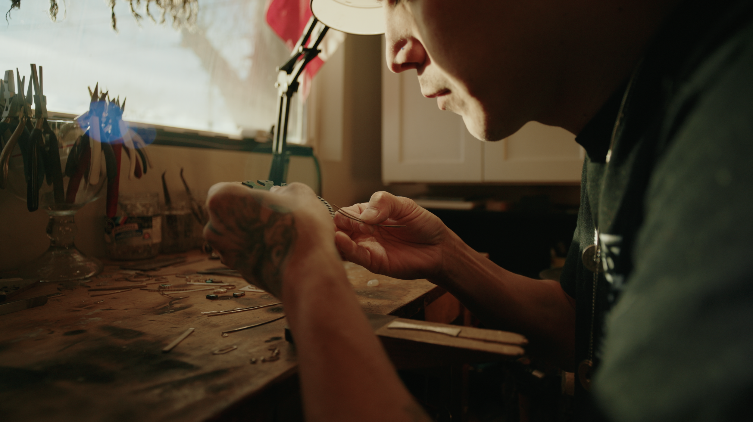 A person with tattoos on their hands working on jewelry at a cluttered workspace, with tools and small items scattered on the table, sunlight coming through a window.