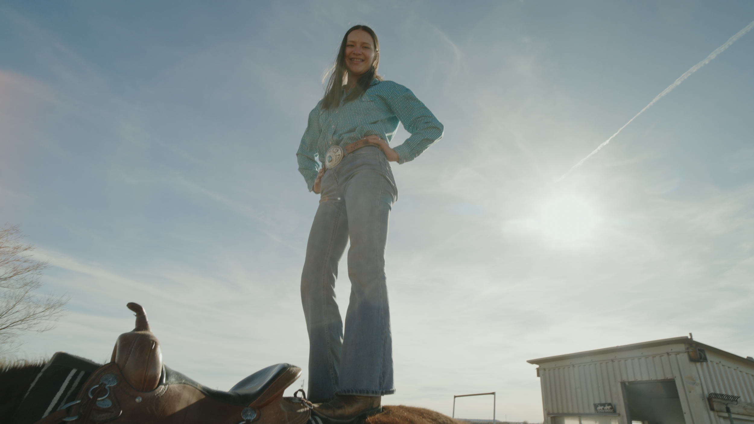 A woman standing on a horse, smiling, with her hand on her hip, wearing western attire including a belt with a large buckle, in an outdoor setting under a blue sky with clouds.
