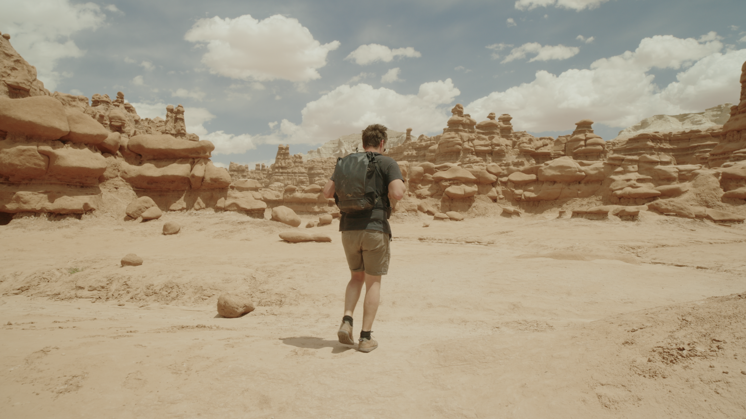A person with a backpack walking through a desert landscape with unique rock formations under a partly cloudy sky.